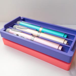 Peut inclure: Un plateau violet à trois compartiments contient trois stylos à insuline de couleur turquoise, crème et rose. Le plateau repose sur une base de couleur corail. Les stylos sont argentés et mesurent 10 unités.
