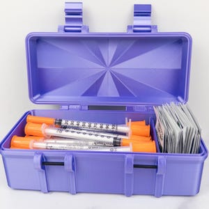 May include: A purple plastic storage case with a hinged lid, containing several clear syringes with orange caps and a stack of small white rectangular packets. The syringes feature black measurement markings ranging from 10 to 100 units.