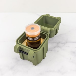 Op de afbeelding: Een olijfgroene, vierkante harde beschermende koffer met een opengeklapt deksel. In de koffer bevindt zich een klein, donker amberkleurig glazen flesje met een doorschijnende plastic dop, dat stevig in een op maat gemaakte inzetstuk rust.