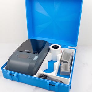 Puede incluir: Un maletín de plástico azul con una impresora de etiquetas gris, un rollo de cinta de etiquetas blanca y dos guías de medición grises. La impresora tiene la marca "NIMBOT". El maletín está abierto, mostrando el contenido.