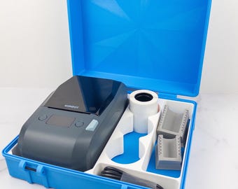 May include: A dark gray Niimbot label printer housed in a bright blue plastic carrying case. The case includes a white paper roll, two gray plastic inserts, and a black charging cable organized within a custom-molded white interior tray.