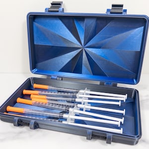 May include: A dark blue rectangular storage case with a geometric starburst pattern on the interior lid. The case is open and contains five clear plastic syringes with orange caps, each featuring black measurement markings along the barrel.