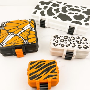 May include: Four rectangular storage cases with hinged lids and latch closures. One large case features a black and white cow print. A square case displays an orange and black monarch butterfly wing pattern. Two smaller cases feature leopard and tiger prints.