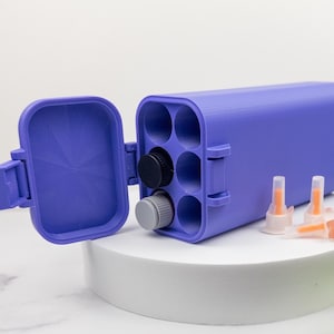 May include: A rectangular purple storage case with a hinged lid and six circular interior compartments. One black cap and one gray cap are inserted into the slots. Two small, clear plastic nozzles with orange tips sit outside the case.