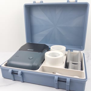 May include: Open blue case containing a grey label maker with the brand name "NIMBOT", two rolls of white labels, and a measuring guide. The case is open, revealing the contents neatly arranged inside.