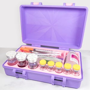 May include: A purple medical kit with a pink interior, open to reveal vials, syringes, and a bottle. The kit has a handle and latches. The vials have silver and yellow caps. The syringes have orange plungers.