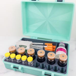 May include: An open, teal-colored medical case containing vials, syringes, and a bottle of sterile water. The case has a foam insert with compartments for the medical supplies. Some vials have yellow caps, while others have tan caps.