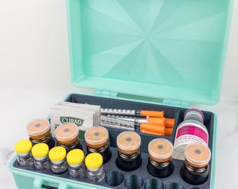 May include: An open, teal-colored medical case containing vials, syringes, and a bottle of sterile water. The case has a foam insert with compartments for the medical supplies. Some vials have yellow caps, while others have tan caps.