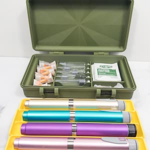 Puede incluir: Un botiquín médico verde oliva abierto con cierres amarillos, que contiene jeringas, agujas y almohadillas con alcohol. Debajo, cuatro plumas de insulina doradas, turquesas, moradas y rosas descansan sobre una bandeja amarilla.