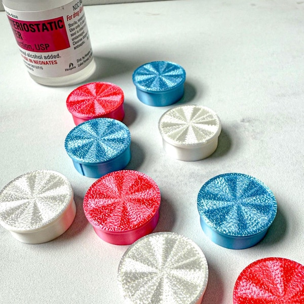 May include: An assortment of colorful, round ear plugs in red, white, and blue. The ear plugs have a radial design on top and are arranged near a bottle with white and red labels. The ear plugs are likely for ear protection.