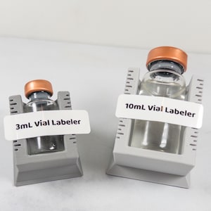 May include: Two clear glass vials with copper-colored caps, labeled as 3mL and 10mL, are displayed in gray holders. The holders have measurement markings. The vials are filled with a clear liquid. The background is a light, neutral color.