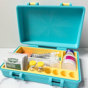 以下が含まれることがあります： 黄色い内装の開いたターコイズ色の医療キット。注射器、アルコールパッド、バイアル、および「Bacteriostatic Water」とラベル付けされたボトルが含まれています。キットには黄色のハンドルと留め金が付いています。