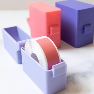 Puede incluir: Tres contenedores pequeños, dos son morados y uno es rosa. Los contenedores morados tienen un rollo de cinta blanca con una etiqueta rosa en el interior.