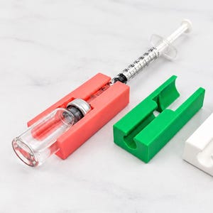 May include: A coral-colored plastic vial holder containing a glass vial and a syringe, positioned next to an empty green plastic vial holder and an empty white plastic vial holder. Each holder features a rectangular base with a central channel.