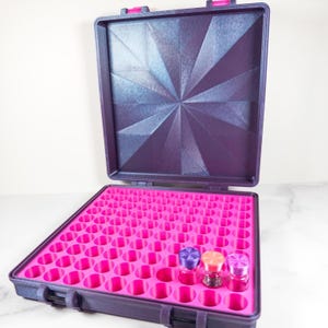 Puede incluir: Un maletín de transporte morado abierto, que revela un interior rosa brillante diseñado para guardar frascos pequeños. La tapa tiene un diseño geométrico. Se ven tres frascos pequeños con tapas de colores dentro del maletín.