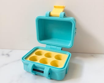 Puede incluir: Una pequeña maleta abierta de color turquesa con un pestillo y un interior amarillos. La maleta tiene un inserto de espuma amarilla con seis recortes circulares. La maleta está diseñada para proteger objetos pequeños.