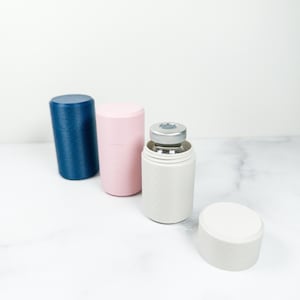 Op de afbeelding: Drie cilindrische houders met een textuur in marineblauw, lichtroze en gebroken wit. De gebroken witte houder is geopend en toont een klein glazen flesje met een zilveren metalen dop, terwijl het bijbehorende deksel er los naast ligt.