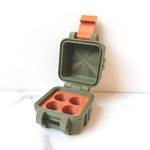 Peut inclure: Un étui carré vert olive ouvert avec un intérieur orange et un fermoir orange assorti. L'intérieur comporte quatre découpes circulaires. L'étui est sur une surface blanche.