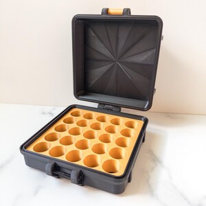 May include: Black carrying case with an open lid. The interior features a yellow insert with multiple circular indentations. The case has a handle and latches. The lid has a geometric design.