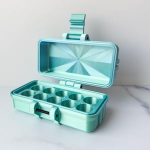 Può includere: Una custodia rettangolare aperta verde menta con coperchio incernierato. L'interno ha otto scomparti circolari. Il coperchio ha un design geometrico. La custodia è realizzata in materiale stampato in 3D e ha una chiusura a scatto.