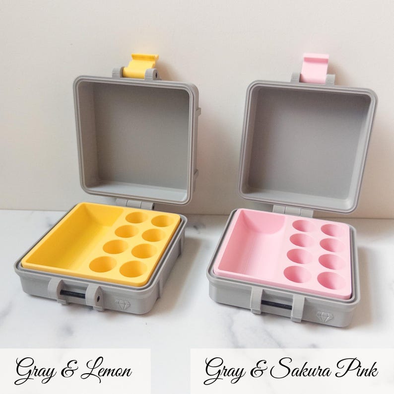 May include: Two open gray cases with yellow and pink interiors. The yellow case has a lemon-colored tray with circular indentations. The pink case has a sakura pink tray with similar indentations. Each case has a gray exterior with a latch.