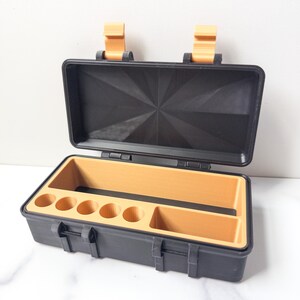 May include: Open black storage case with a textured lid and a tan interior. The case has a series of circular compartments and a rectangular compartment. The case has two tan latches and two tan hinges.