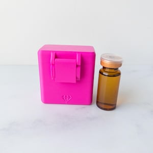 Op de afbeelding: Een felroze vierkante opbergdoos met een scharnierend deksel en een klein ruitvormig logo in reliëf op de voorzijde, naast een klein amberkleurig glazen flesje met een metalen dop gevuld met een doorschijnende bruine vloeistof.
