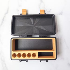 May include: An open, rectangular black and orange storage box. The lid is black with a geometric pattern. The interior has a black compartment and an orange section with six circular holes and a rectangular space. The box has orange clasps.