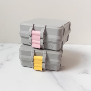 May include: Two gray, stackable storage containers with pink and yellow clasps. Each container has a diamond-shaped logo on the front. The containers are designed with a rugged, industrial aesthetic.