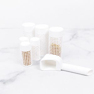 以下が含まれることがあります： 茶色の内部が見える穴あきメッシュパターンの白い円筒形容器5個と、長方形のボウルと長い柄を持つ白いプラスチック製のスクープが、平らな面に配置されています。