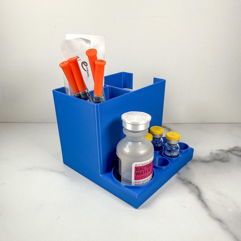 May include: A vibrant blue organizer with multiple compartments. It holds orange syringes, small vials with yellow caps, and a bottle labeled "Bacter Water for Injection." The organizer is designed to hold medical supplies.