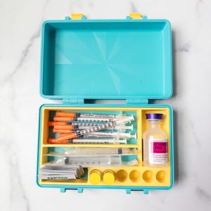 K&ouml;nnte beinhalten: Ein t&uuml;rkisfarbener Kunststoff-Aufbewahrungskoffer mit gelbem Inneneinsatz, der mehrere Spritzen mit orangefarbenen Kappen, eine kleine Glasflasche mit der Aufschrift Bacteriostatic Water, einzeln verpacktes medizinisches Zubeh&ouml;r und kleine F&auml;cher enth&auml;lt.