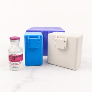 May include: A clear glass vial of Bacteriostatic Water for injection with a pink cap and label, alongside a small blue rectangular plastic storage case, a larger white rectangular plastic storage case, and a purple rectangular plastic storage case.