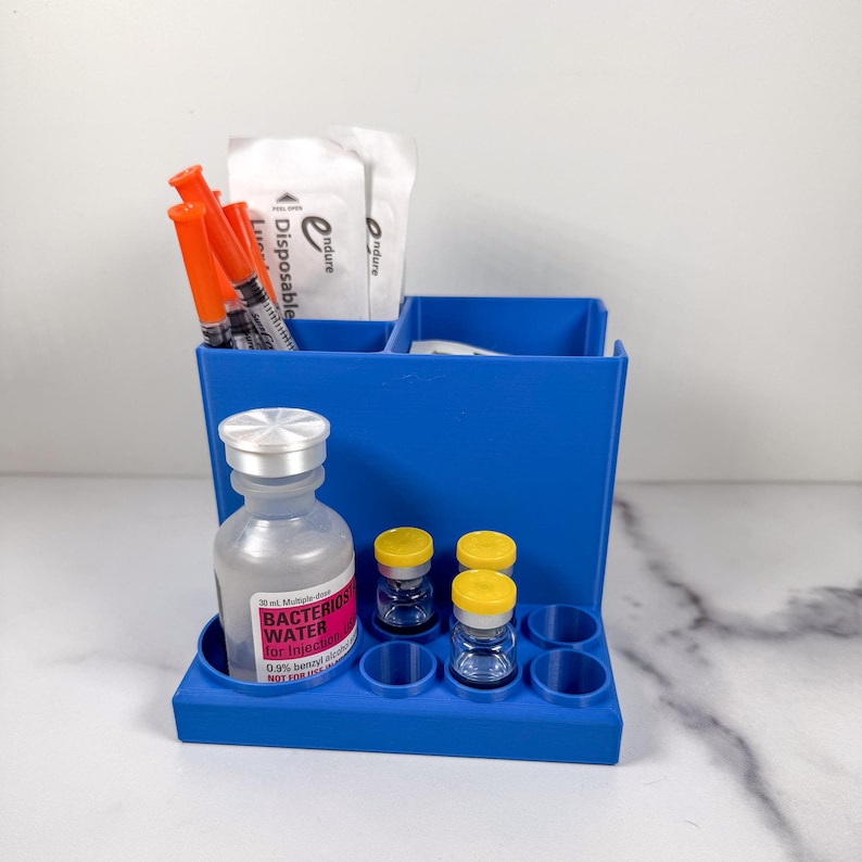 May include: A blue plastic organizer holds medical supplies. It contains orange syringes, small vials with yellow caps, and a bottle labeled "Bacterios Water for Injection." The organizer has multiple compartments and circular holders.