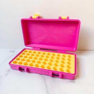 Puede incluir: Maleta de plástico rosa abierta con interior amarillo. La maleta tiene un asa y cierres. El interior tiene múltiples hendiduras circulares. La maleta está diseñada para guardar objetos pequeños.