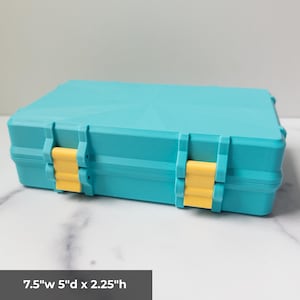 K&ouml;nnte beinhalten: Eine rechteckige t&uuml;rkisfarbene Aufbewahrungsbox mit zwei gelben Kunststoffverschl&uuml;ssen an der Vorderseite. Die Box hat ein geometrisches Sternmuster auf dem Deckel und misst 19,05 cm Breite, 12,7 cm Tiefe und 5,72 cm H&ouml;he laut Aufdruck.