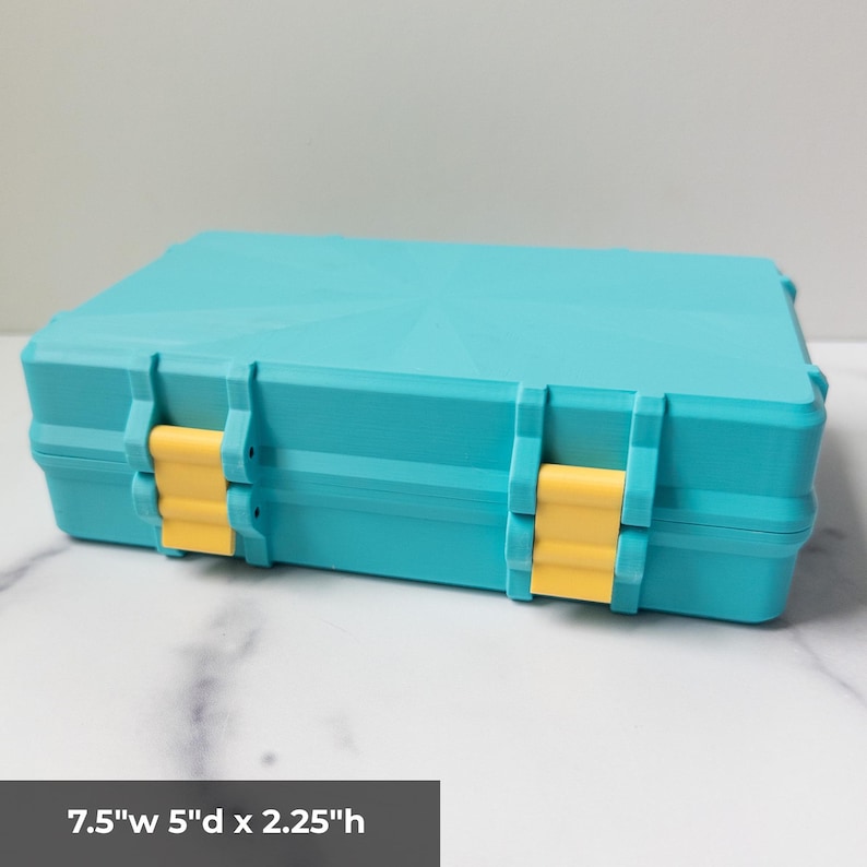 K&ouml;nnte beinhalten: Eine rechteckige t&uuml;rkisfarbene Aufbewahrungsbox mit zwei gelben Kunststoffverschl&uuml;ssen an der Vorderseite. Die Box hat ein geometrisches Sternmuster auf dem Deckel und misst 19,05 cm Breite, 12,7 cm Tiefe und 5,72 cm H&ouml;he laut Aufdruck.