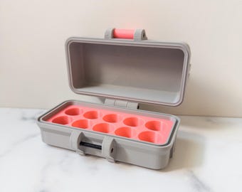 Puede incluir: Una caja rectangular gris abierta, con interior rosa y asa rosa. El interior tiene compartimentos circulares. La caja está sobre una superficie de mármol blanco.