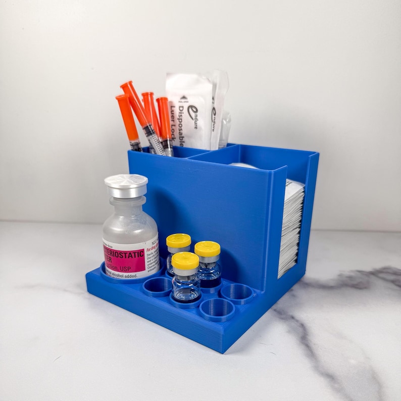 May include: A blue organizer with multiple compartments. It holds syringes, vials, and medical supplies. The organizer has a tray with circular holders for small bottles. The background is a white surface.