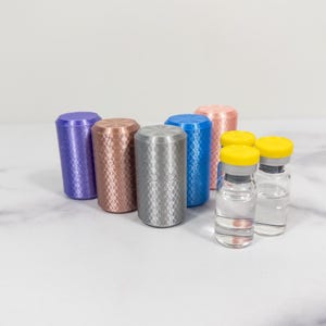 Op de afbeelding: Een verzameling kleurrijke, getextureerde cilindrische objecten en kleine glazen flesjes met gele doppen. De cilinders zijn in tinten paars, koper, zilver, blauw en roze. De flesjes zijn gevuld met een heldere vloeistof.