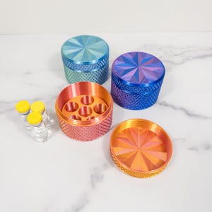 May include: A selection of brightly coloured, cylindrical containers with textured sides and starburst-patterned lids. The containers are in shades of blue, purple, orange, and pink. Small glass vials with yellow caps are also present.