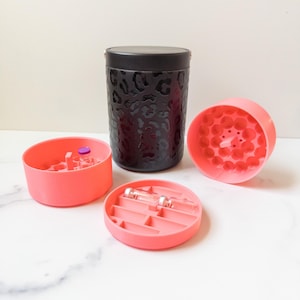 May include: A black container with a leopard print pattern, a black lid, and gold accents. Three coral-coloured, circular containers are open, revealing vials. The containers are designed to hold small items.