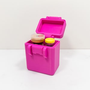 May include: A bright pink, rectangular storage box with an open hinged lid. Inside the container are two small glass vials, one with a metallic gold cap and the other with a bright yellow cap, held securely within the upright, open-top case.