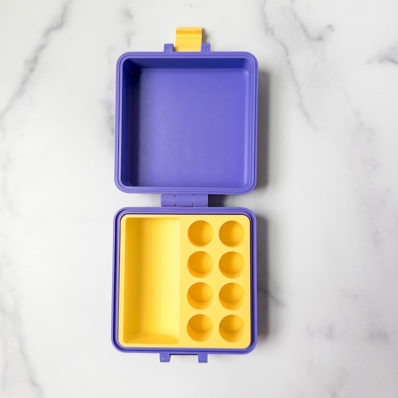 May include: An open, square, purple and yellow container with a yellow latch. The interior is yellow with eight circular indentations. The top half is purple. The container is on a white marble surface.
