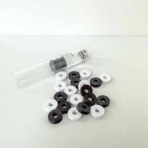 May include: A clear glass vial with a silver cap and a dark gray stopper, filled with a clear liquid. Scattered around are small, round, black and white decorative beads. The beads have a hole in the center.