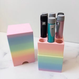 May include: A pastel rainbow gradient storage container with a matching lid, featuring a square base with six circular slots. Three cylindrical pens in black, teal, and pink are inserted into the slots, each with a grey cap and a small window.