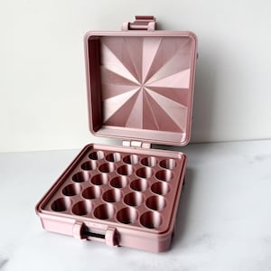May include: A pink, open, small carrying case with a geometric design on the lid. The case has a tray with 24 circular indentations. The case is made of a solid material and has a handle and latches.