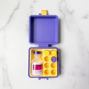 May include: An open, purple and yellow medical kit containing a bottle with a label, yellow discs, and a purple disc. The bottle has a white label with red and black text. The kit is on a white marble surface.