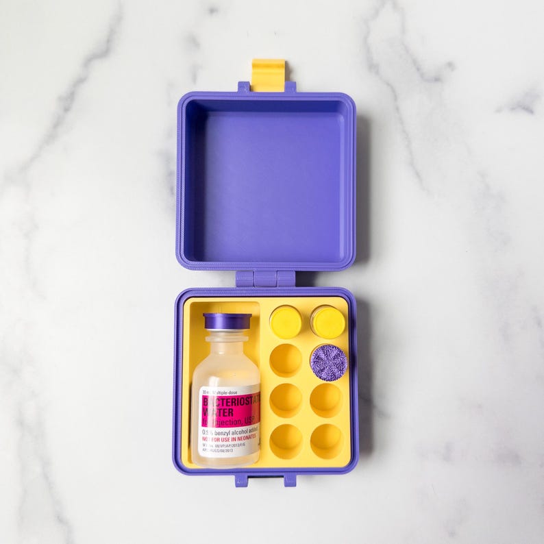 May include: An open, purple and yellow medical kit containing a bottle with a label, yellow discs, and a purple disc. The bottle has a white label with red and black text. The kit is on a white marble surface.