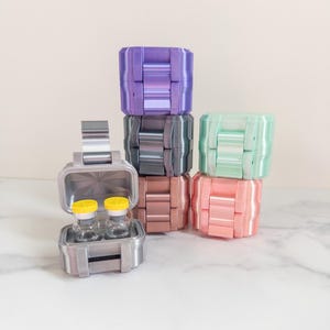 May include: A selection of 3D-printed boxes in various colours, including purple, grey, rose, mint green, and pink. One box is open, displaying two small glass vials with yellow caps. The boxes are stacked, featuring a modern, geometric design.